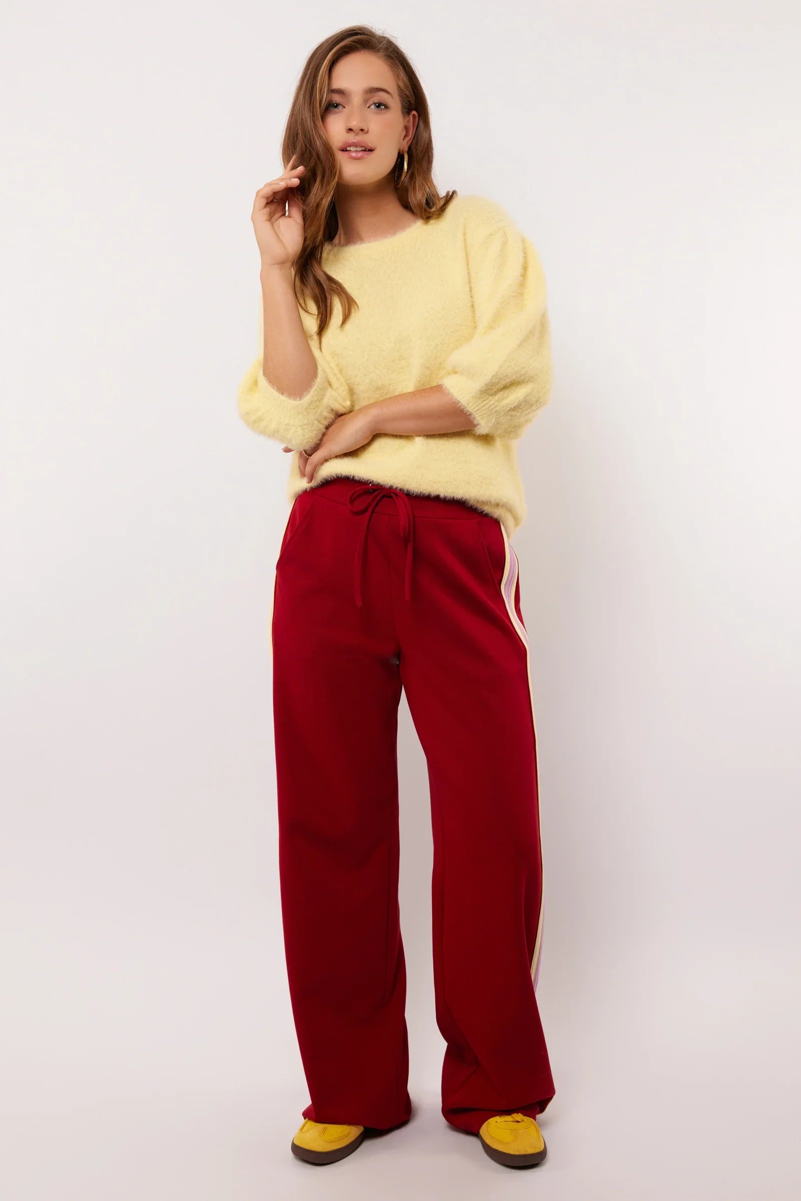 Model draagt rode Fluresk Jorina pants met pastelgele en lila zijstrepen, gecombineerd met een zachte gele trui.