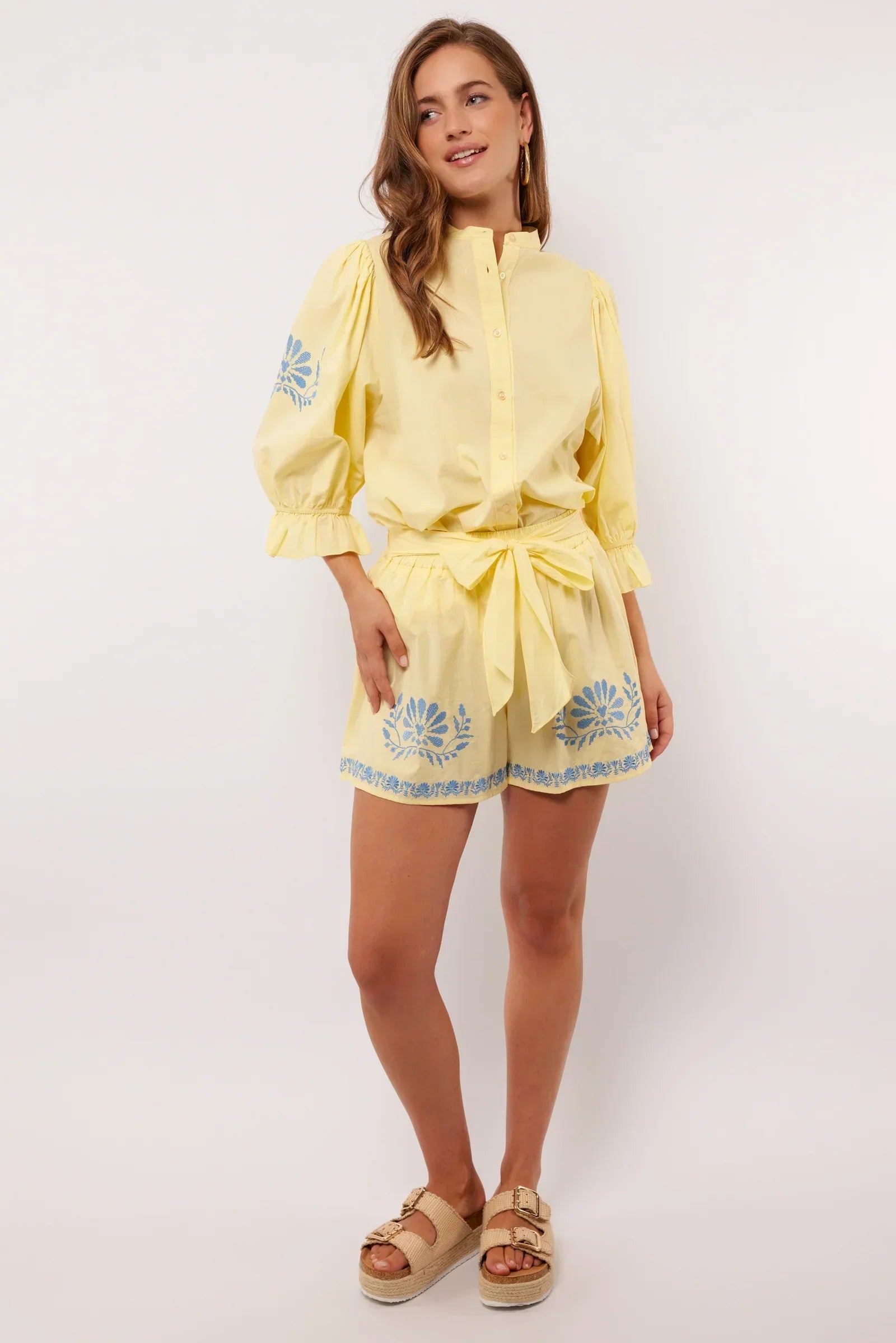 Vrouw draagt gele Fluresk Saine short met blauwe bloemenprint en bijpassende blouse, zomerse outfit met sandalen.