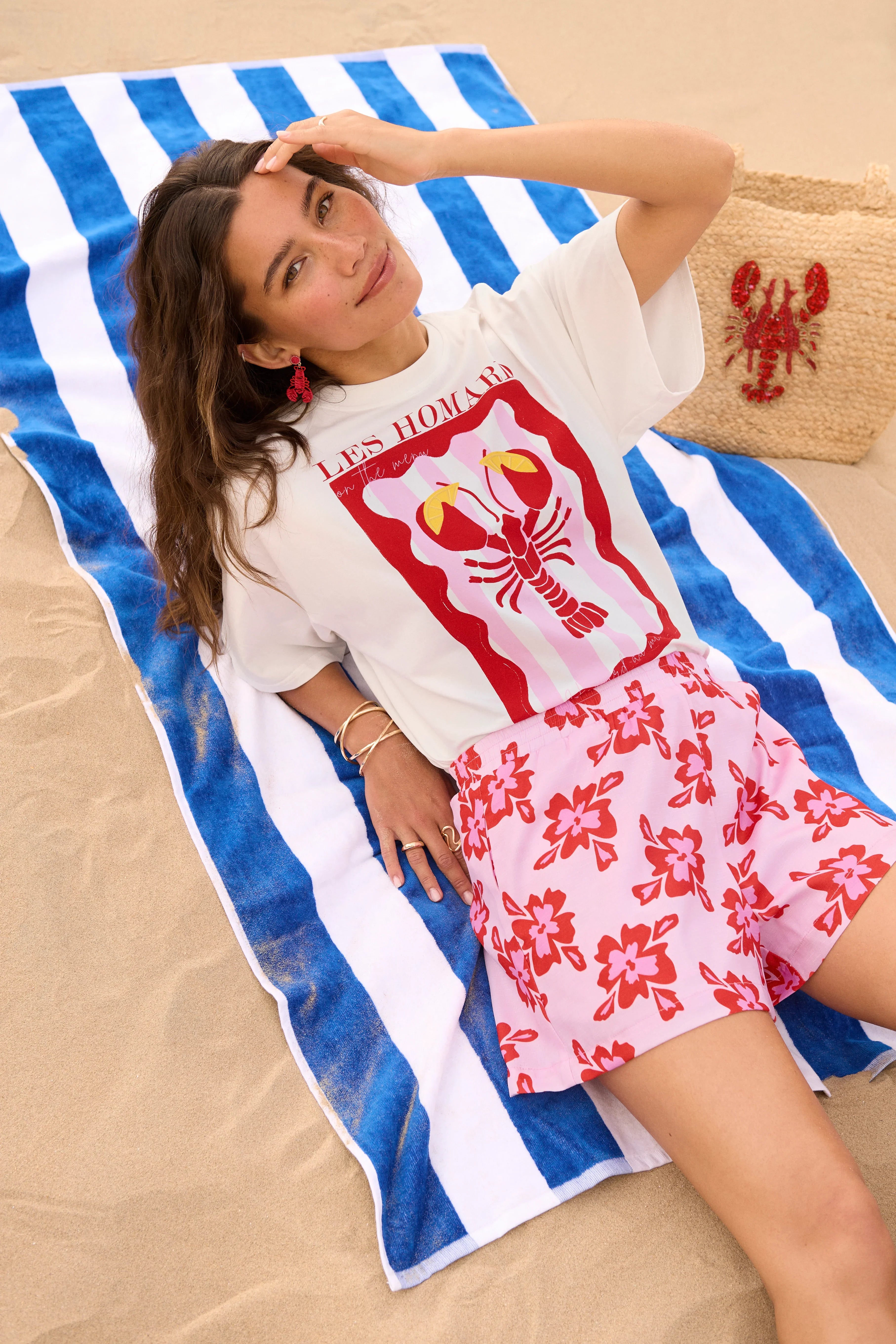 Model draagt het witte Ydence Lobster T-shirt met rode kreeftprint, liggend op een blauw-wit gestreept strandlaken.