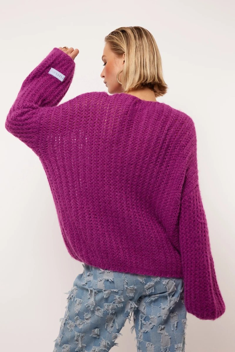 Model draagt de Florin Cardigan van Fluresk in een levendige paarse kleur. De achterkant van de cardigan wordt benadrukt, met een losse pasvorm en zachte gebreide textuur. De cardigan is gecombineerd met lichtblauwe jeans met een ruige afwerking.