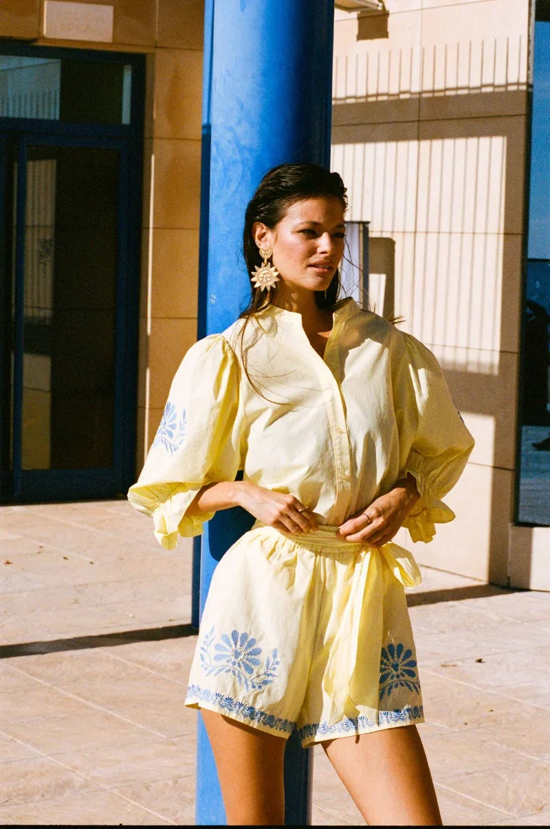 Model draagt de gele Marlowe blouse en Saine shorts van Fluresk met blauwe bloemenborduursels, in de zon bij een modern gebouw.