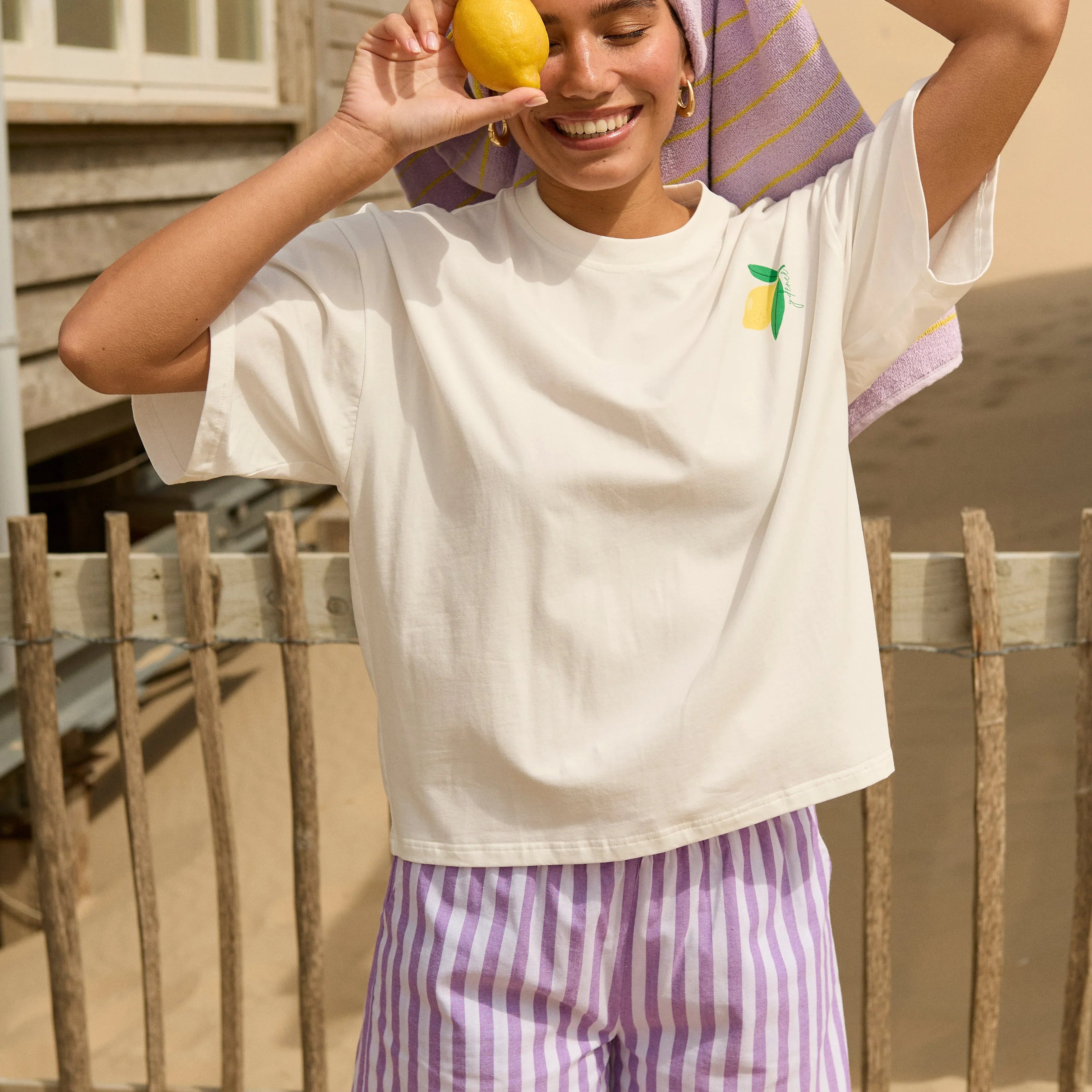 Model draagt Ydence mima short met lila-witte strepen, gecombineerd met een wit t-shirt met citroen bedrukking en lila handoek op het strand. Zomerse outfit uit de Ydence zomercollectie. 
