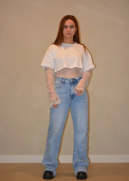 Model draagt een lichtblauwe jeans met een rechte pasvorm en subtiele wassing, gestyled met een witte cropped top en kanten details voor een trendy look.