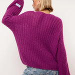 Model draagt de Florin Cardigan van Fluresk in een levendige paarse kleur. De achterkant van de cardigan wordt benadrukt, met een losse pasvorm en zachte gebreide textuur. De cardigan is gecombineerd met lichtblauwe jeans met een ruige afwerking.