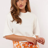 Model draagt het witte Fluresk Brix T-shirt in een casual outfit met een oranje bloemenrok en gouden oorbellen.