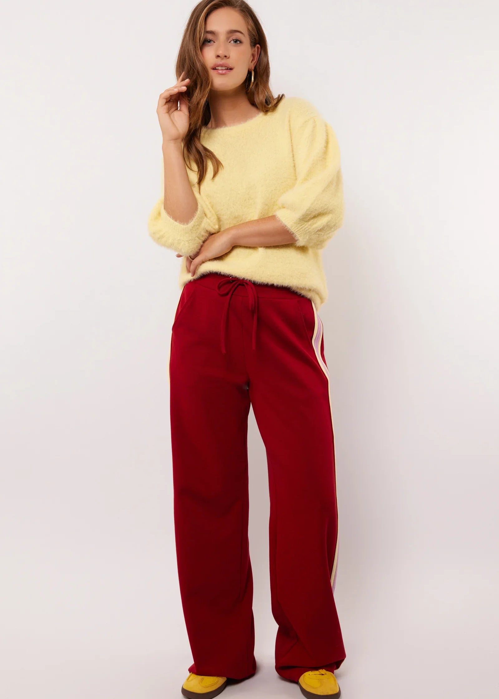 Model draagt rode Fluresk Jorina pants met pastelgele en lila zijstrepen, gecombineerd met een zachte gele trui.