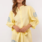 Model draagt gele Fluresk Marlowe blouse met bijpassende short en blauwe borduursels.