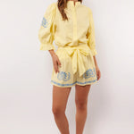 Vrouw draagt gele Fluresk Saine short met blauwe bloemenprint en bijpassende blouse, zomerse outfit met sandalen.