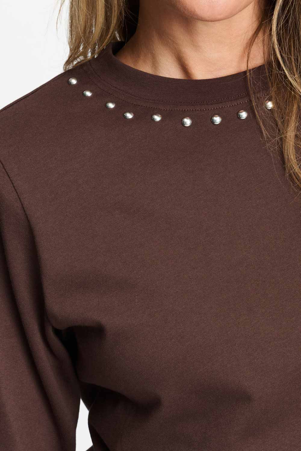 Detailfoto van de zilverkleurige studs rond de halslijn van het bruine t-shirt.