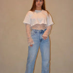 Model draagt een witte cropped top gecombineerd met een kanten top eronder en een lichtblauwe jeans met een casual pasvorm, gestyled met zwarte schoenen.