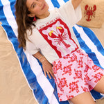 Model draagt het witte Ydence Lobster T-shirt met rode kreeftprint, liggend op een blauw-wit gestreept strandlaken.