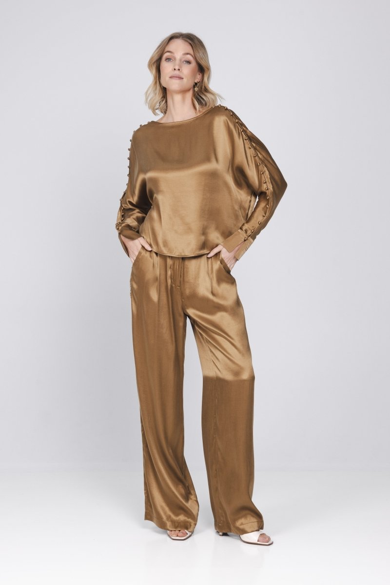 Een model toont een complete, glanzende bronskleurige set, bestaande uit een top | Accentil Mira Pants Mokka | Jo&Co Club