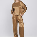 Een model toont een complete, glanzende bronskleurige set, bestaande uit een top | Accentil Mira Pants Mokka | Jo&Co Club