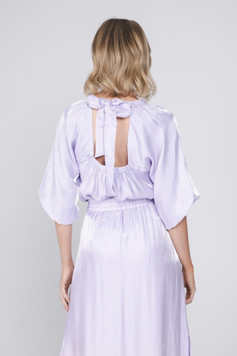 Een model, van de rug gefotografeerd, draagt een glanzende, lila jurk | Accentil Sere Blouse Lila | Jo&Co Club
