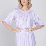 Een model met blond, halflang golvend haar staat gefotografeerd tegen een effen | Accentil Sere Blouse Lila | Jo&Co Club
