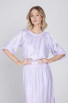 Een model met blond, halflang golvend haar staat gefotografeerd tegen een effen | Accentil Sere Blouse Lila | Jo&Co Club