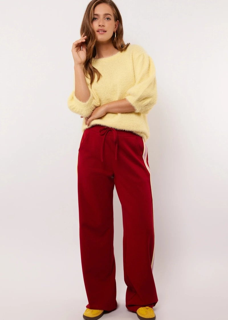 Model draagt rode Fluresk Jorina pants met pastelgele en lila zijstrepen, gecombineerd met een zachte gele trui.