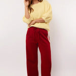 Model draagt rode Fluresk Jorina pants met pastelgele en lila zijstrepen, gecombineerd met een zachte gele trui.