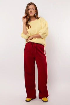 Model draagt rode Fluresk Jorina pants met pastelgele en lila zijstrepen, gecombineerd met een zachte gele trui.