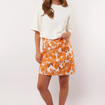 Model showt de Fluresk Mindy Skirt met opvallende oranje bloemenprint, gestyled met een wit T-shirt en retro sneakers.
