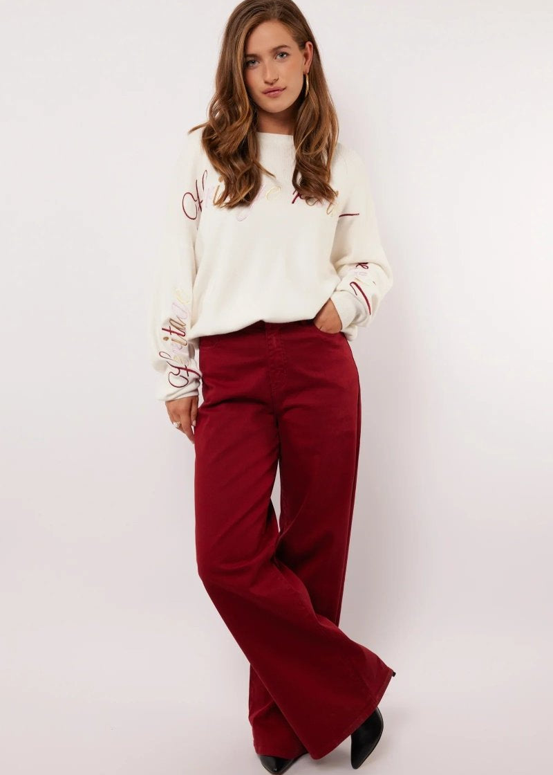 Model draagt de Fluresk Verina Pants in bordeauxrood met witte trui en zwarte laarzen.