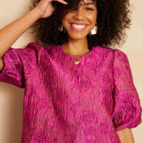 Close-up van een lachend model met volumineus krullend haar dat een fuchsia jacquard top | Lofty Manner Gianna | Jo&Co Club