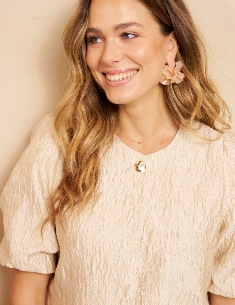Een model met lang, golvend blond haar draagt een crèmekleurige top met een opvallende | Lofty Manner Gianna | Jo&Co Club