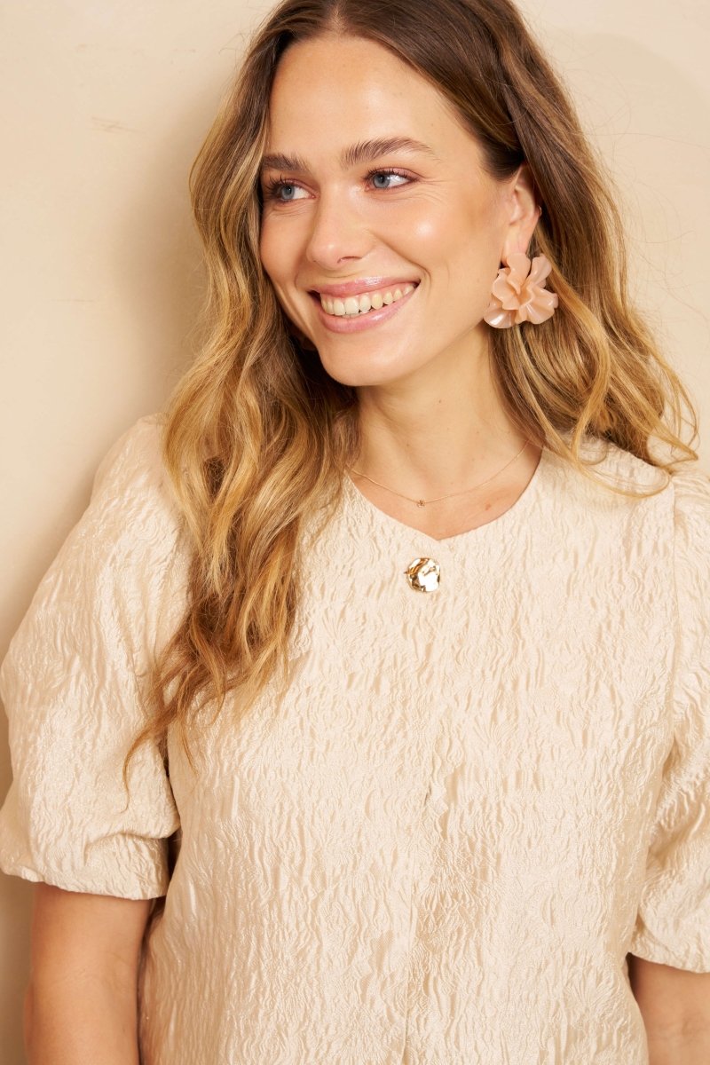 Een model met lang, golvend blond haar draagt een crèmekleurige top met een opvallende | Lofty Manner Gianna | Jo&Co Club