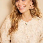 Een model met lang, golvend blond haar draagt een crèmekleurige top met een opvallende | Lofty Manner Gianna | Jo&Co Club