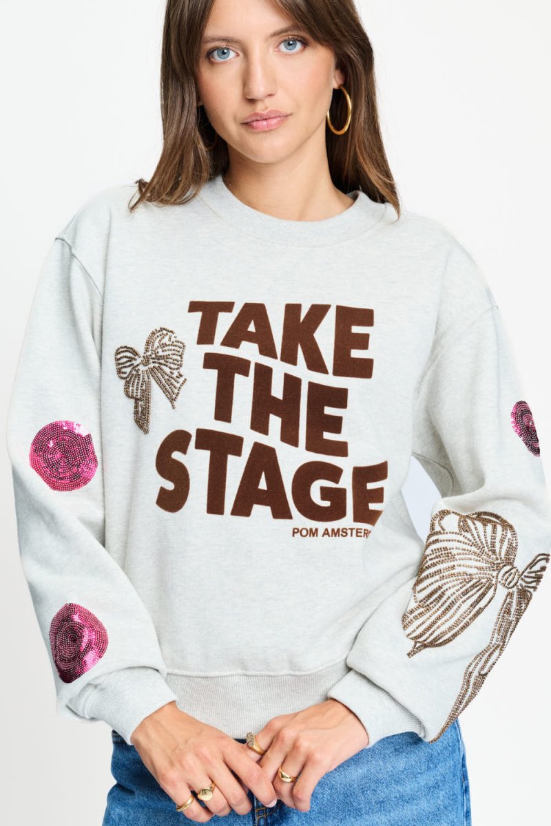 Grijze sweater met ronde ribhals en pailletstrikken op de mouwen, van dichtbij gefotografeerd.