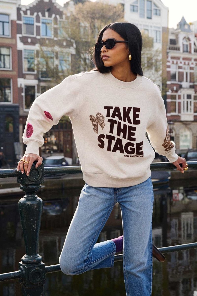 Model draagt de grijze sweater met pailletstrikken op de mouwen aan een Amsterdamse gracht.