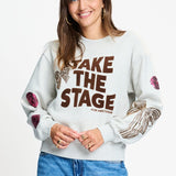 Model draagt de lichtgrijze sweater Sascha van POM Amsterdam met bruine TAKE THE STAGE tekst.