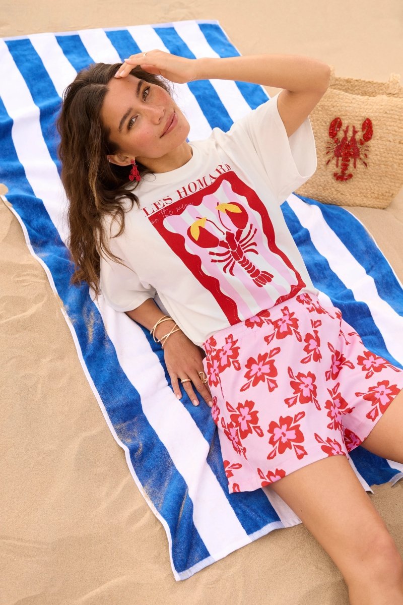 Model draagt het witte Ydence Lobster T-shirt met rode kreeftprint, liggend op een blauw-wit gestreept strandlaken.