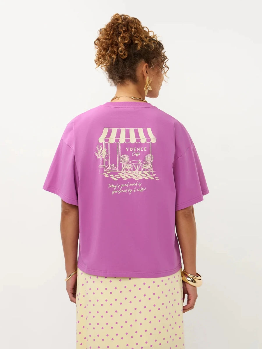 Een model, van achteren gefotografeerd, draagt een oversized magenta T-shirt tegen een witte | Ydence cafe | Jo&Co Club